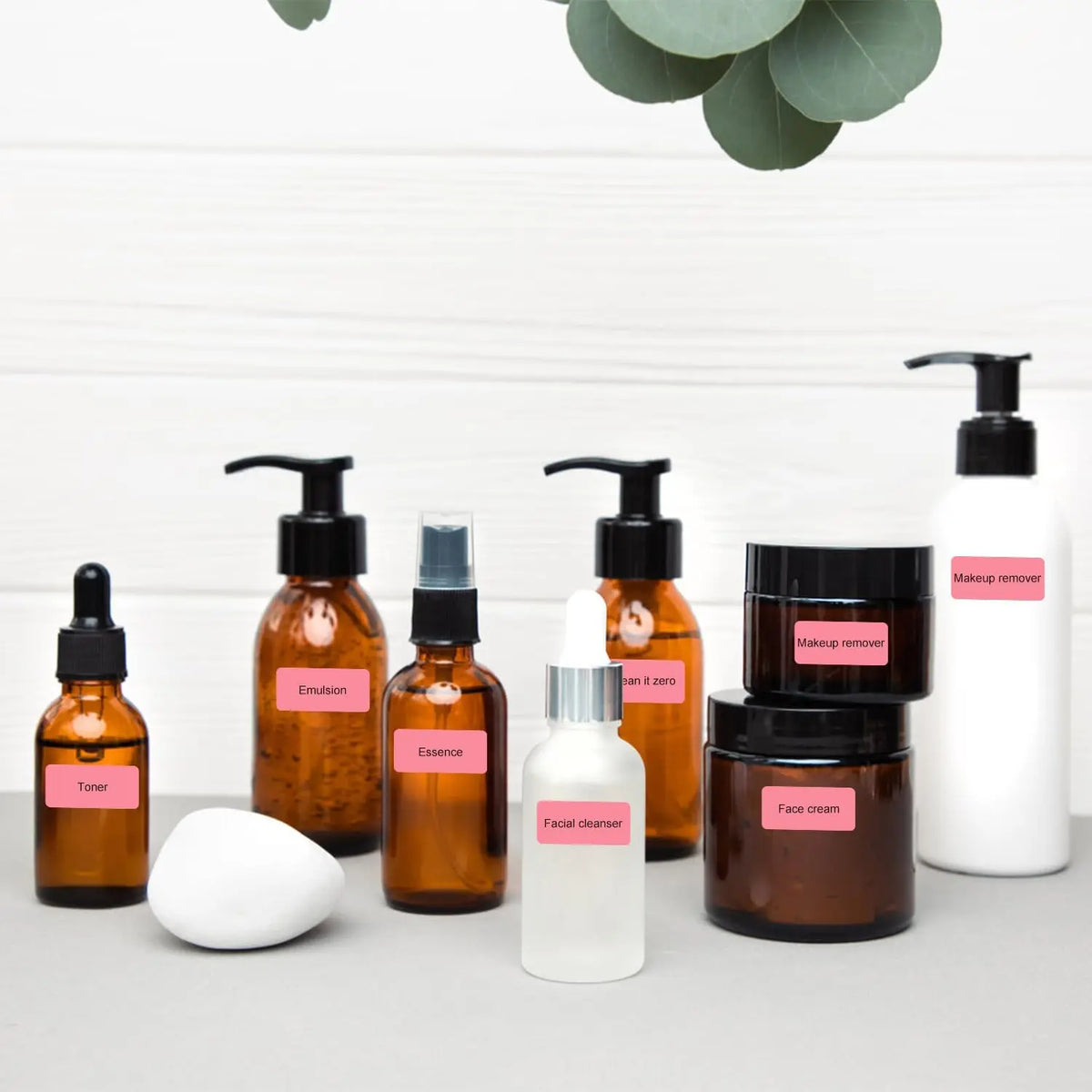 Flacons de cosmétiques ambrés et pots foncés posés sur un plan clair, identifiés chacun par une étiquette rose