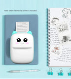 Imprimante à oreilles menthe posée sur un carnet bleu, imprimant un texte, à côté d'un carnet à spirale illustré et d'un stylo